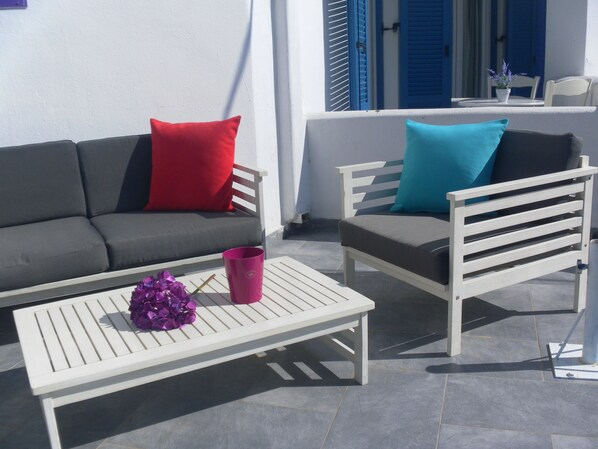 A terrace with a white table, a grey sofa with red and blue cushions, and a white chair with a blue cushion.