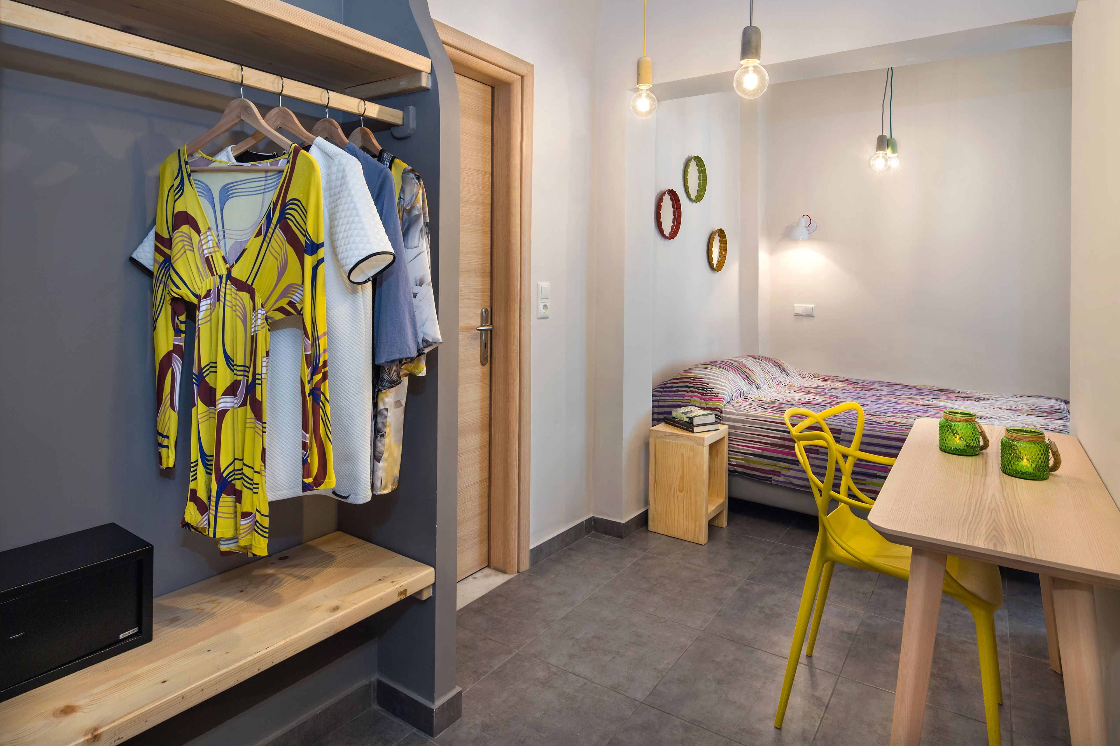 A bedroom with a bed, a bedside table with two green candles, a yellow chair, a wooden bench, and a clothing rack with various garments.