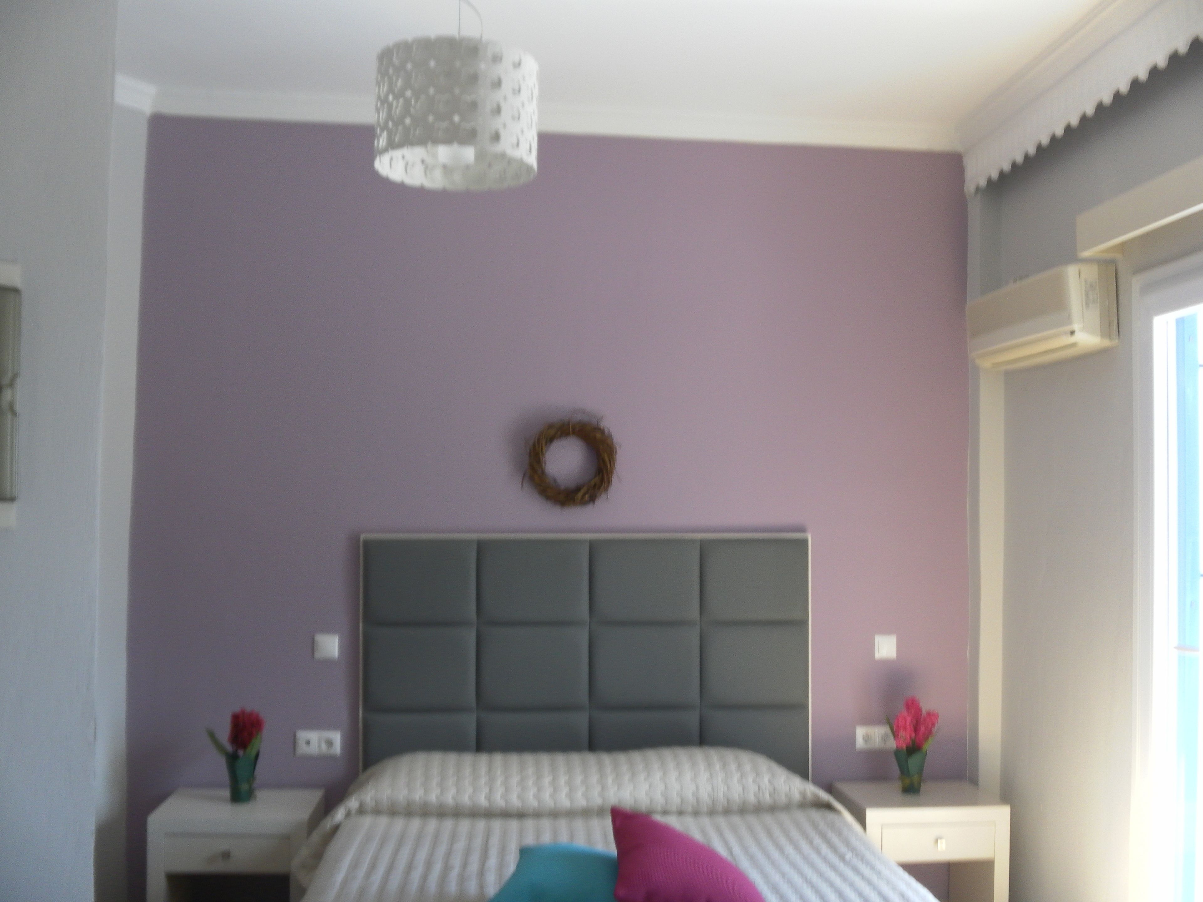 A bedroom with a bed, a headboard, a wall-mounted wreath, and a nightstand with a vase of flowers.