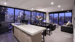 A modern kitchen with a large island, dining area with chairs and a sofa, and large windows offering a city view. - AKOM AT Docklands (Docklands)