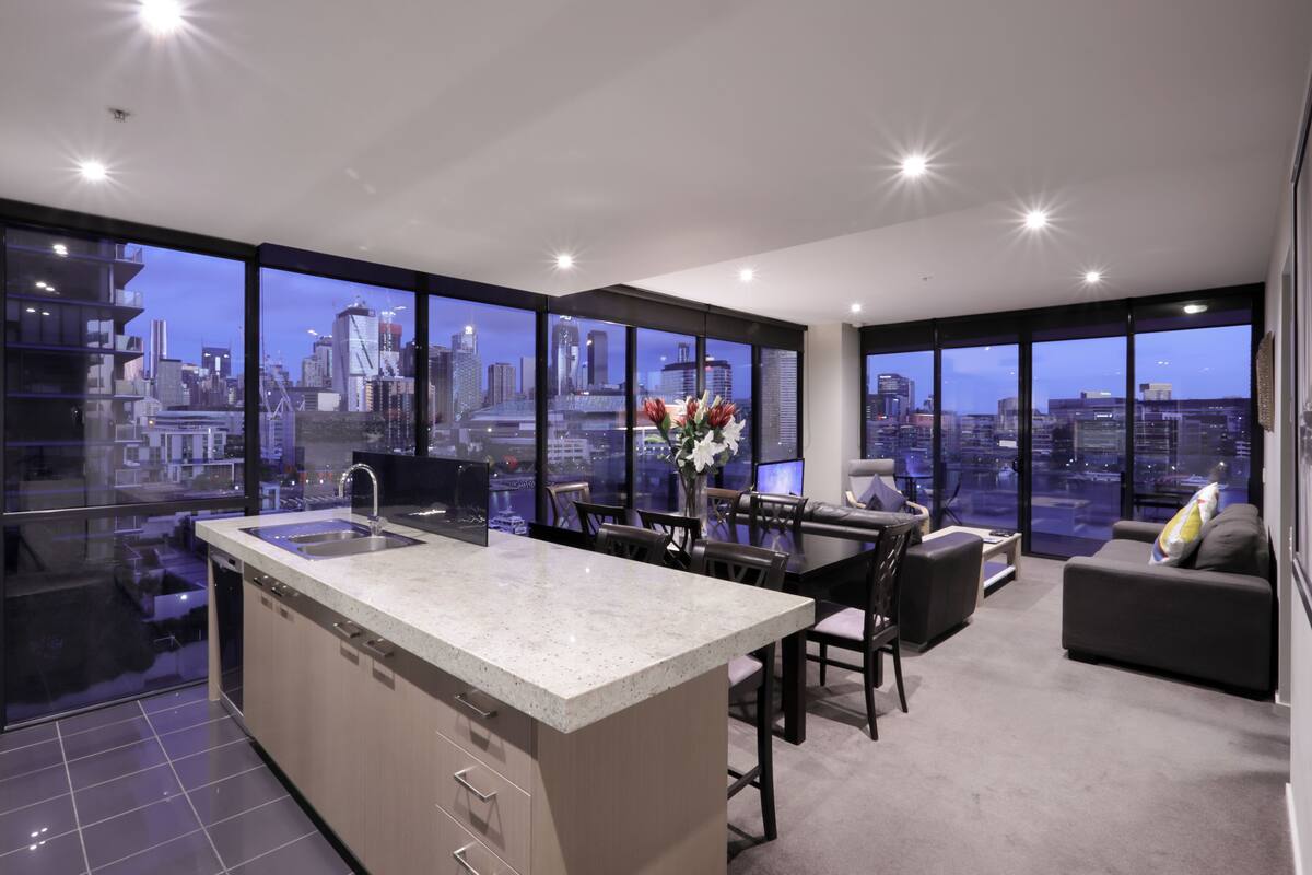 A modern kitchen with a large island, dining area with chairs and a sofa, and large windows offering a city view.