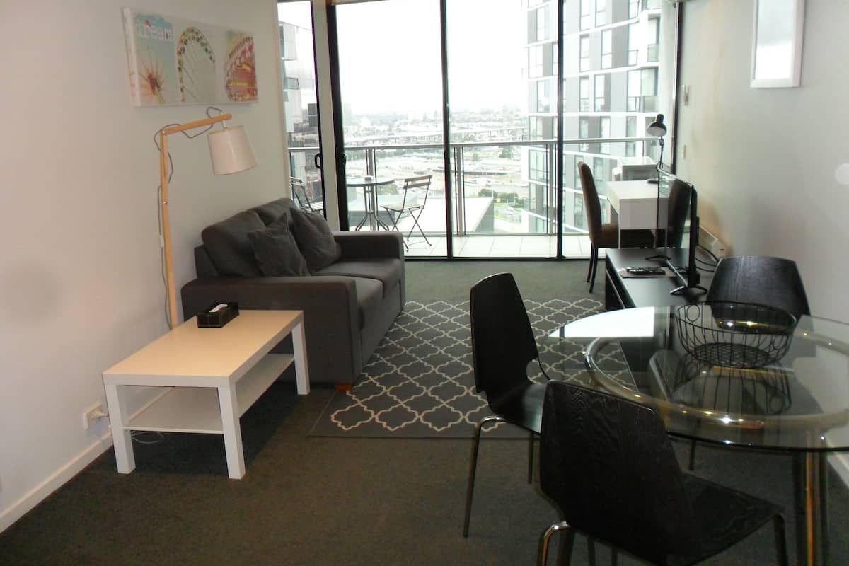 A modern living room with a sofa, a small table, a glass dining table with chairs, and a large window offering a city view.