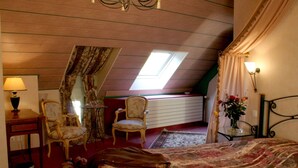 Un dormitorio con techo inclinado, una cama, dos sillas, una mesita, una lámpara y una ventana con cortinas.