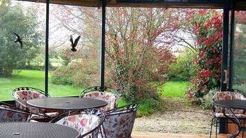 An outdoor covered patio with metal tables and chairs, a chandelier, and a view of a garden with trees and bushes.