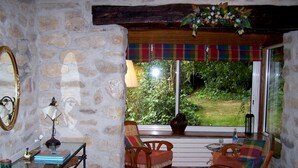 Una habitación acogedora con paredes de piedra, una mesa de madera, dos sillas, una mesita con lámpara y vistas al verdor exterior.