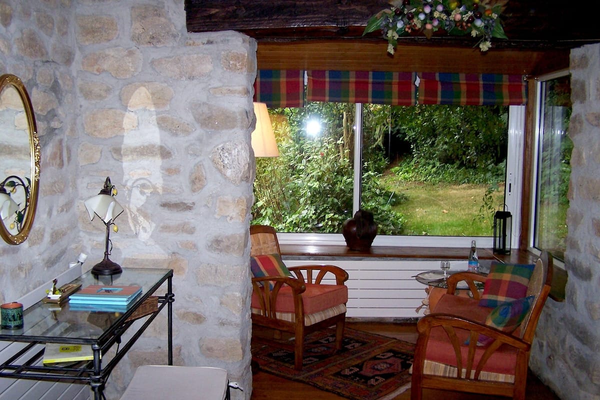 A cozy room with stone walls, a wooden table, two chairs, a small table with a lamp, and a view of greenery outside.