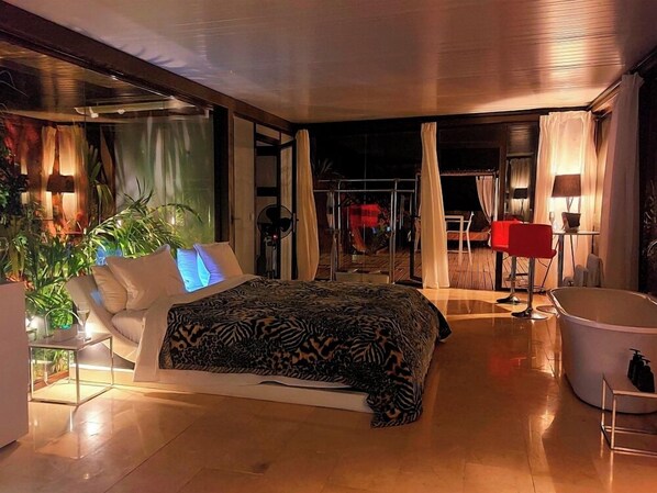 Un dormitorio moderno con una cama grande, una zona de estar con una silla roja, una bañera y un área de patio con paredes de vidrio.