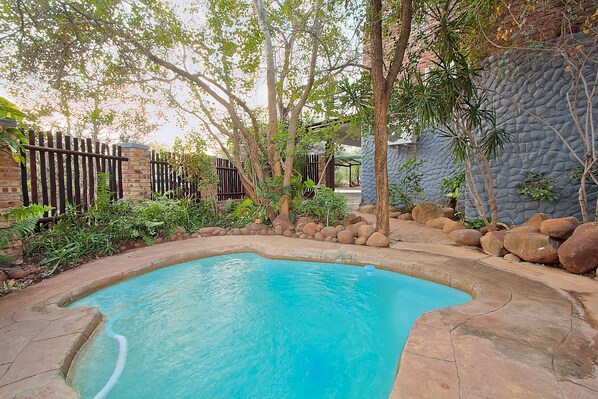 A swimming pool surrounded by a stone wall, a wooden fence, and various plants. - Royale Marlothi Safari Lodge (Nkomazi)
