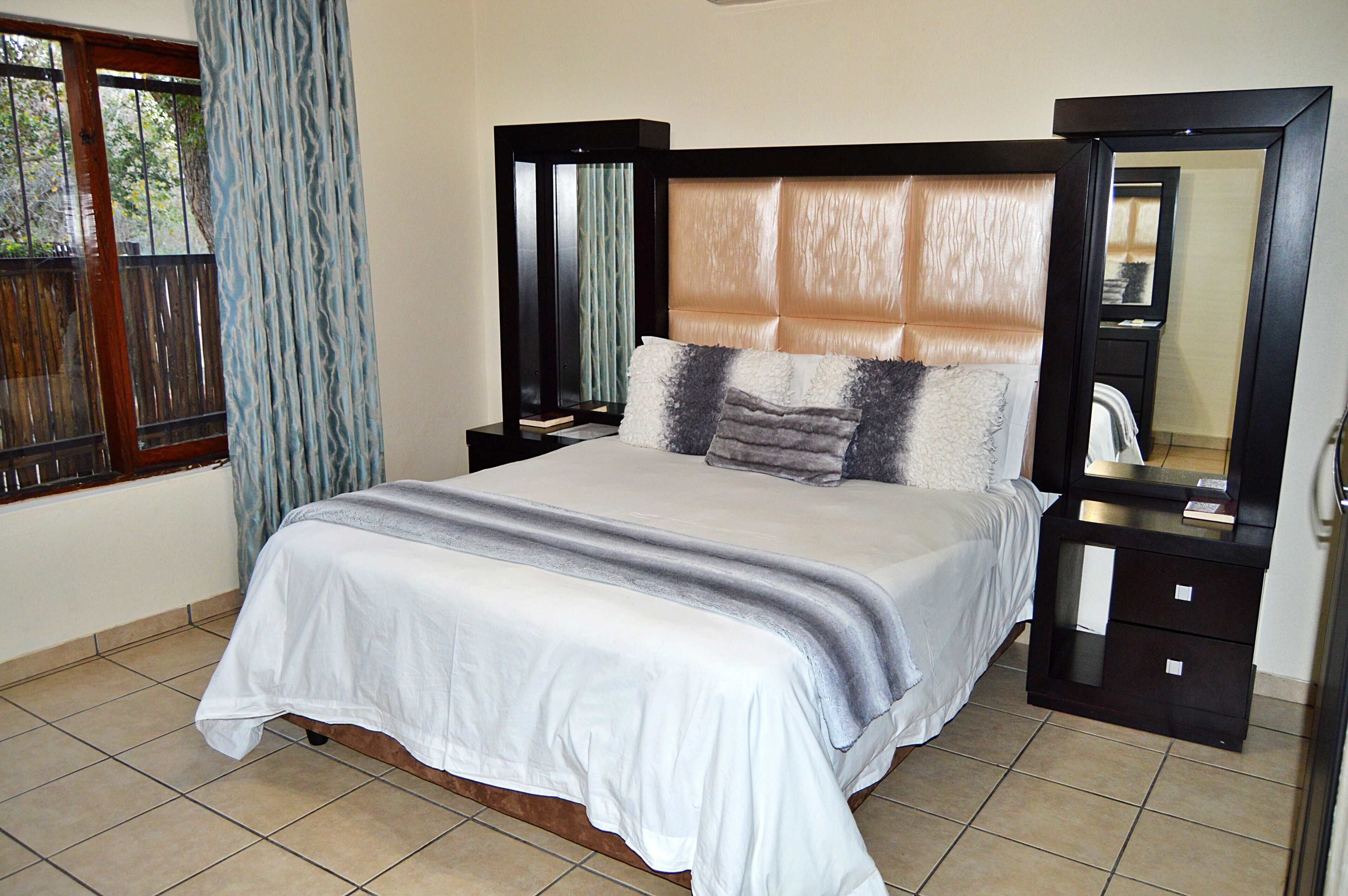 A neatly made bed with a white and gray striped blanket, a headboard with a light brown panel, and a nightstand with a mirror and a small drawer.