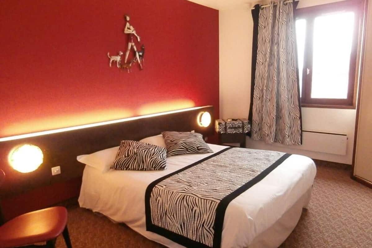 A hotel room with a bed, a chair, a headboard with lights, a zebra-patterned bedspread, a wall decoration of a person walking a dog, and a window with curtains.
