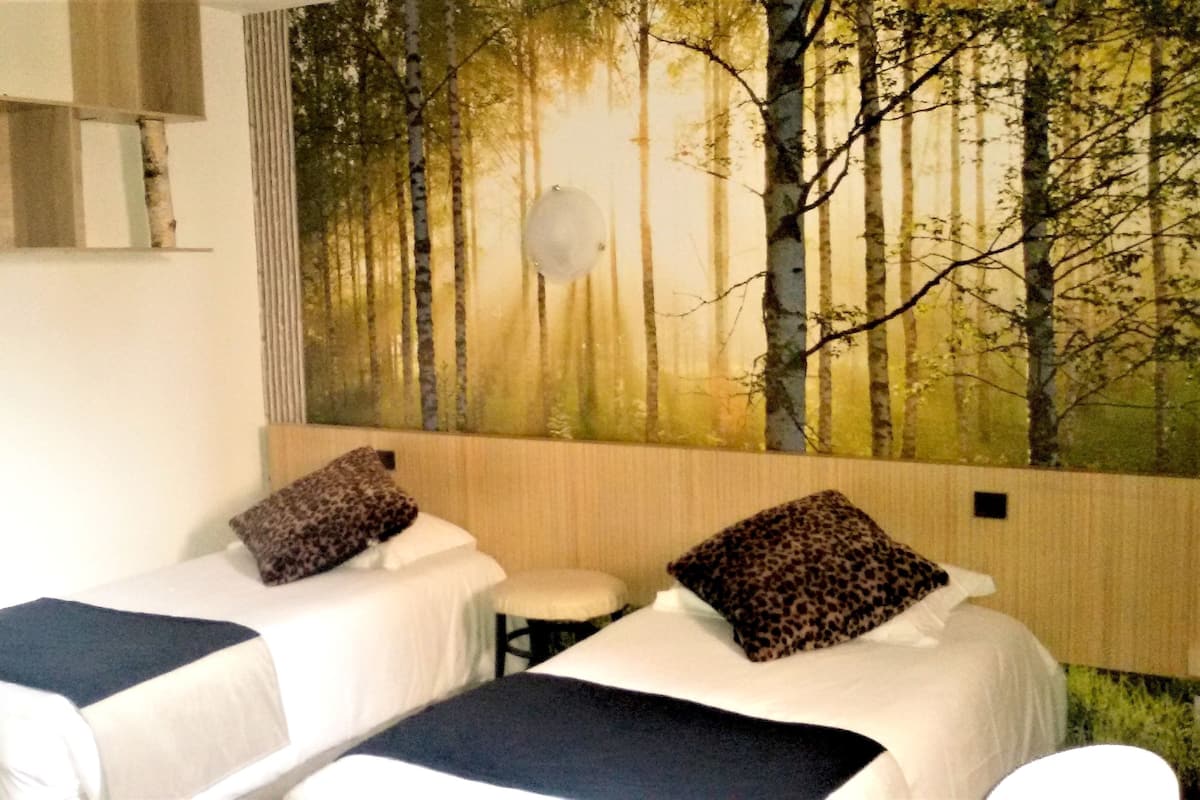 A room with two beds, each with a pillow and a blanket. There is a large forest mural on the wall behind the beds.