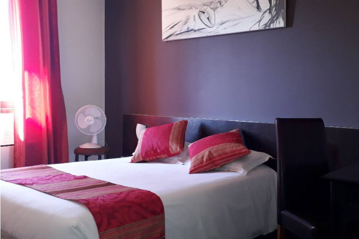 A hotel room with a large bed, red and blue pillows, a white bedspread, a fan, and a painting on the wall.