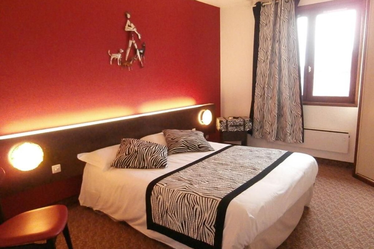 A hotel room with a bed, a chair, a headboard with lights, a zebra-patterned bedspread, a wall decoration of a person walking a dog, and a window with curtains.