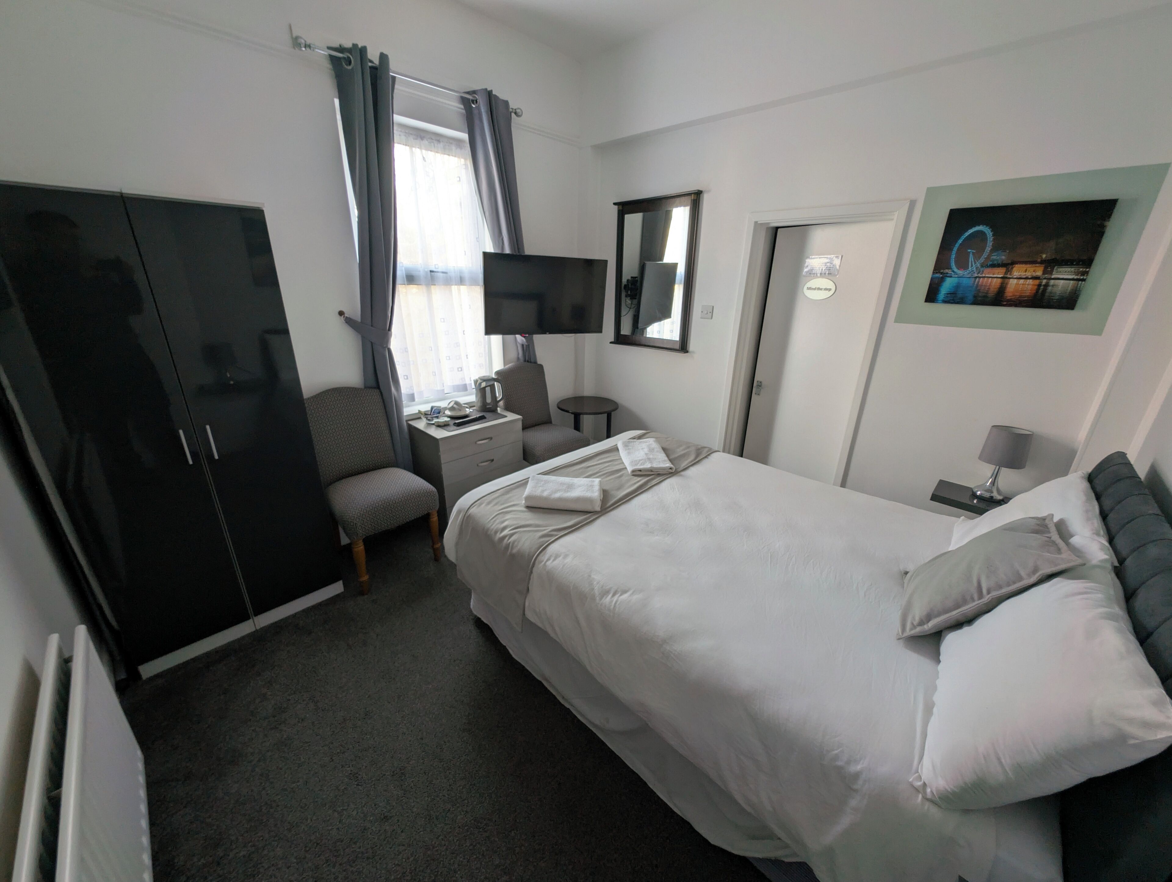 A hotel room with a bed, a desk with a computer, a chair, a wardrobe, a television, and a painting on the wall.