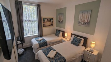 A hotel room with two beds, a window with curtains, a painting of sailboats, and a lamp on the nightstand.