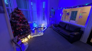 A living room with a Christmas tree, blue ambient lighting, a black leather sofa, and a window with blinds. - Cecil Court Hotel (Bournemouth)