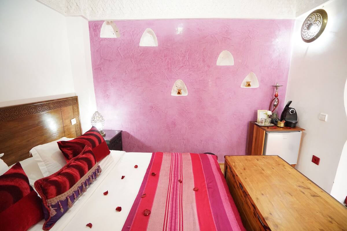 A bedroom with a bed, a wooden nightstand, a small table, and a wall-mounted shelf with a hookah.
