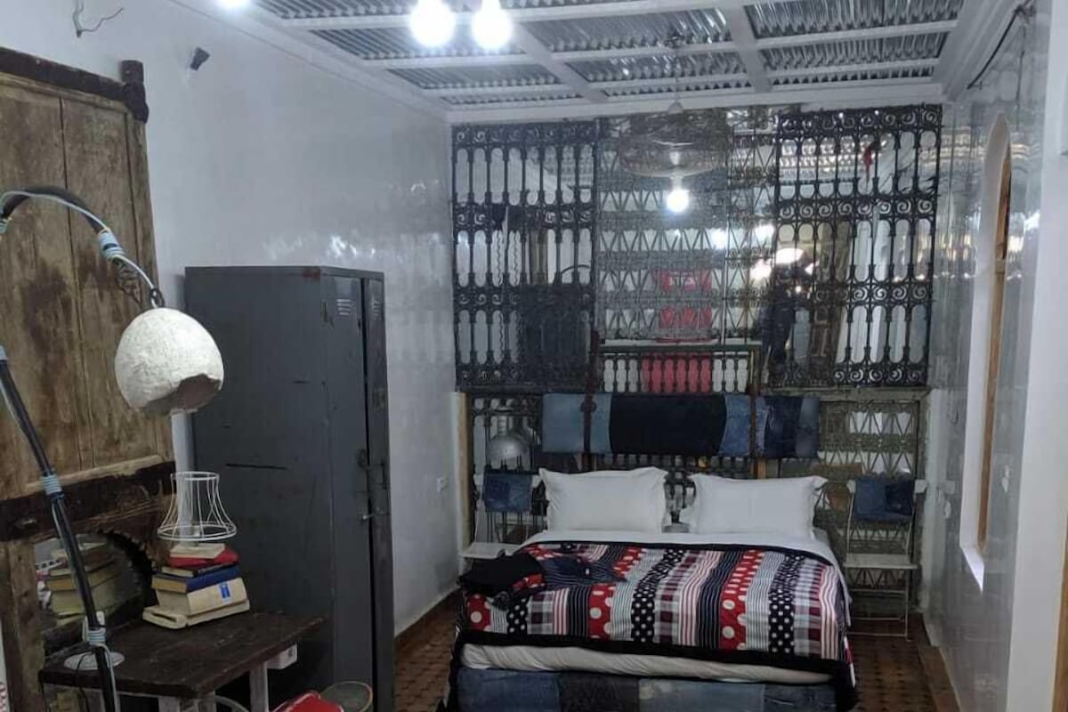 A compact room with a bed, a desk, and a chair. There is a bookshelf and a window with metal bars.