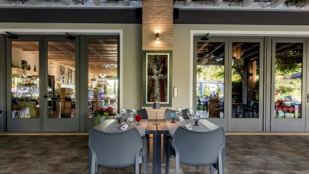 An outdoor dining area with a table set for two, surrounded by grey chairs, and a view of a restaurant through glass doors.