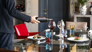 A man pouring wine into a glass at a dining table with a red chair and a decorative vase in the background.