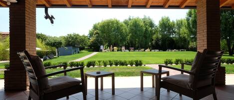 An outdoor patio with two chairs and a small table, a brick pillar, and a view of a green lawn and trees.
