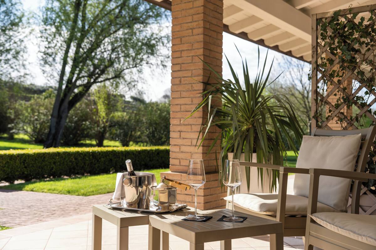 An outdoor patio with a table set for two, a chair, and a view of a garden.