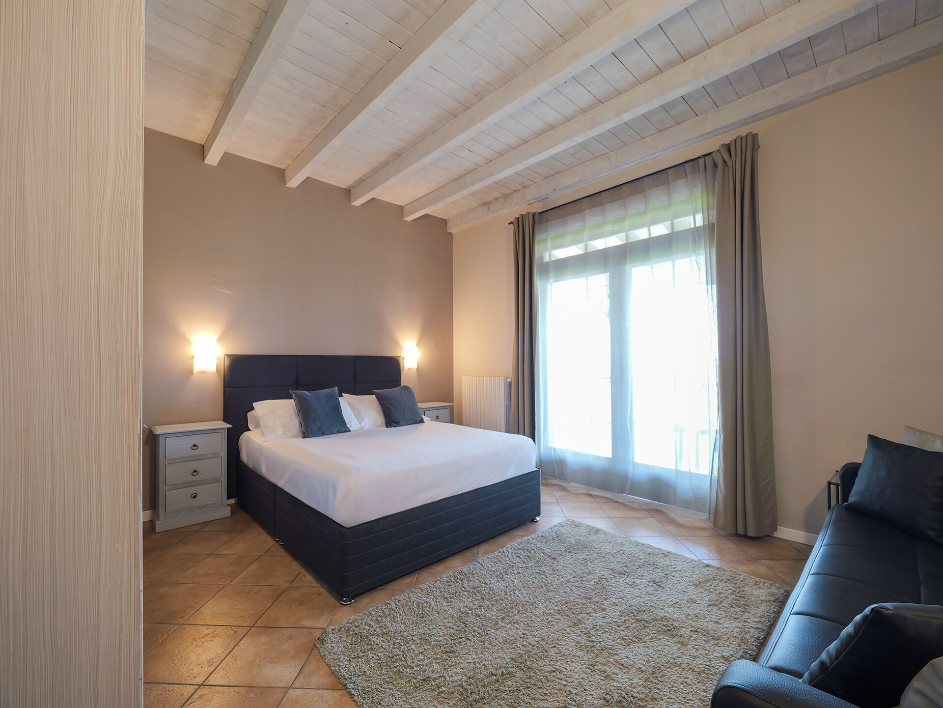 A bedroom with a bed, bedside tables, a nightstand, a sofa, a large window with curtains, and a wooden ceiling.