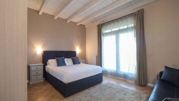 A bedroom with a bed, bedside tables, a nightstand, a sofa, a large window with curtains, and a wooden ceiling.