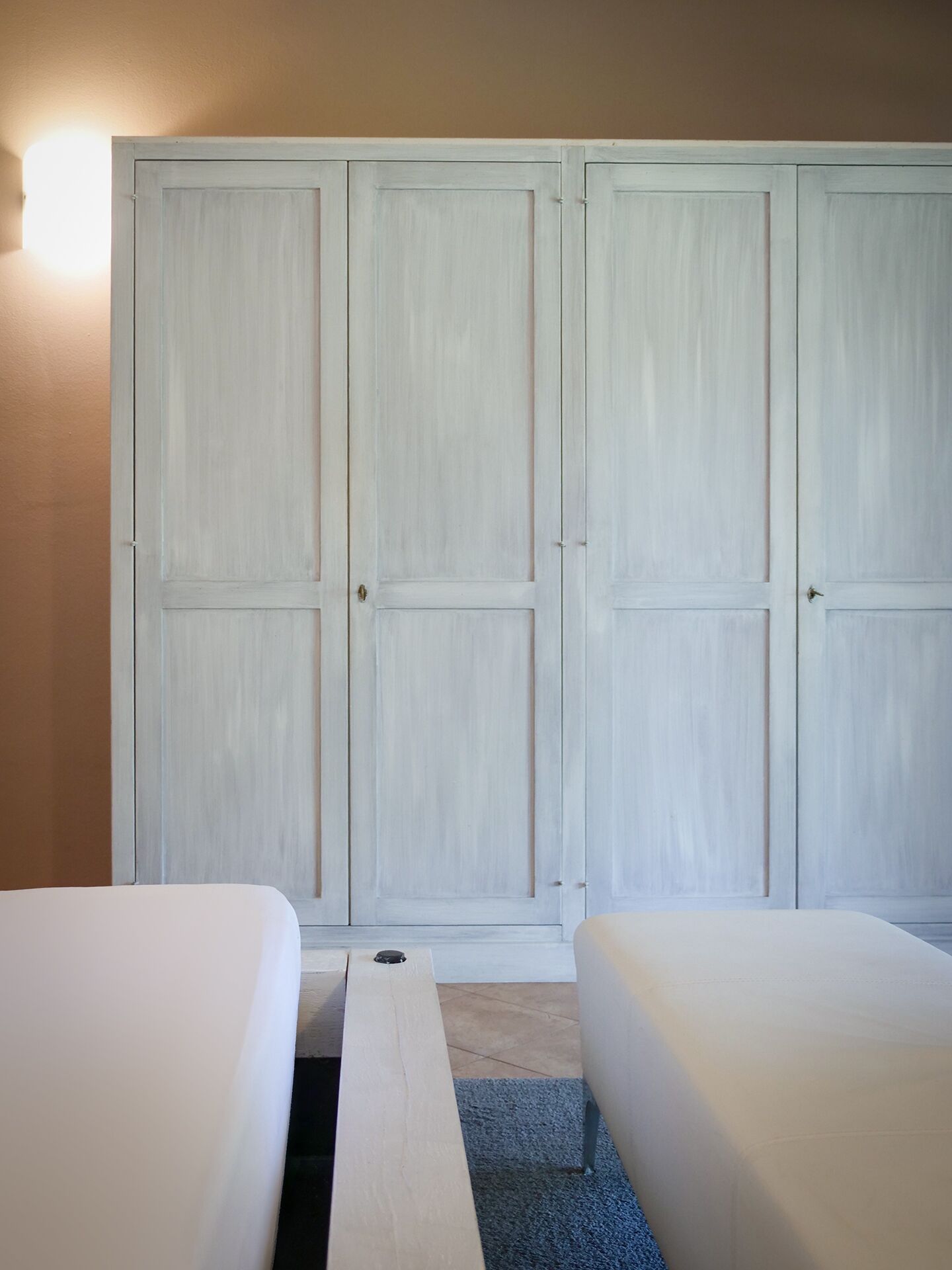 A room with two beds, a large white wardrobe, and a light fixture on the wall.