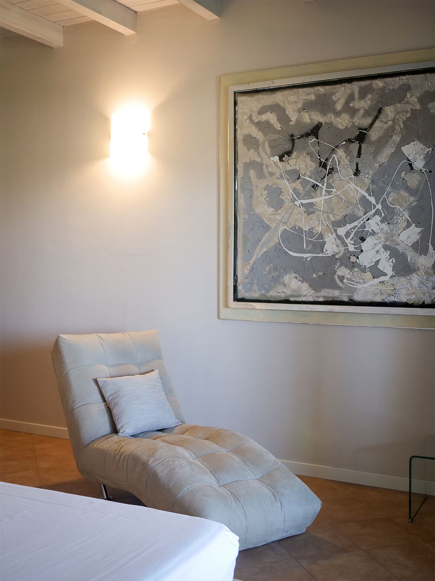 Una habitación con una silla acolchada, una cama y un cuadro abstracto grande en la pared.