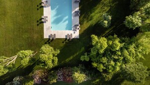 An aerial view of a rectangular pool with a surrounding deck, surrounded by lush greenery and trees.