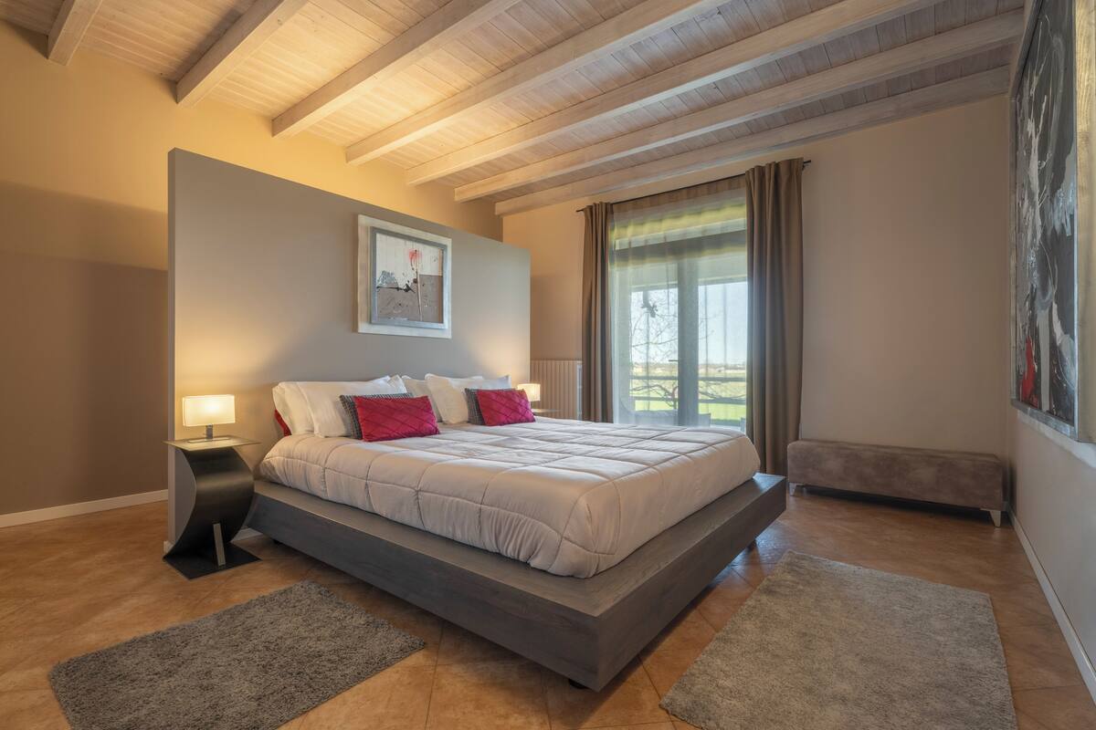 A bedroom with a large bed, a bedside table, a nightstand, a window with curtains, and a radiator.