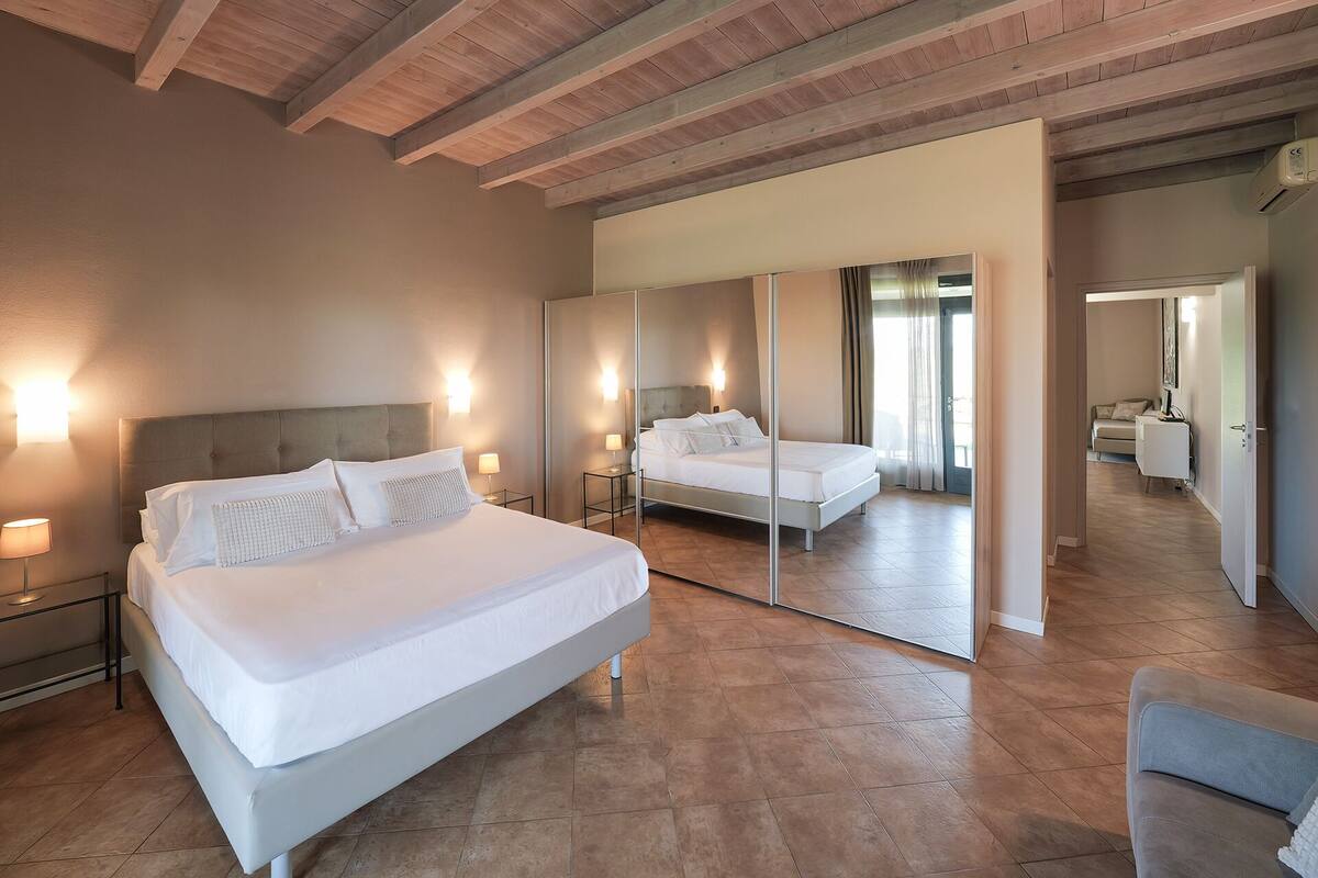 A hotel room with a bed, bedside tables, a mirror, and a view of another room.