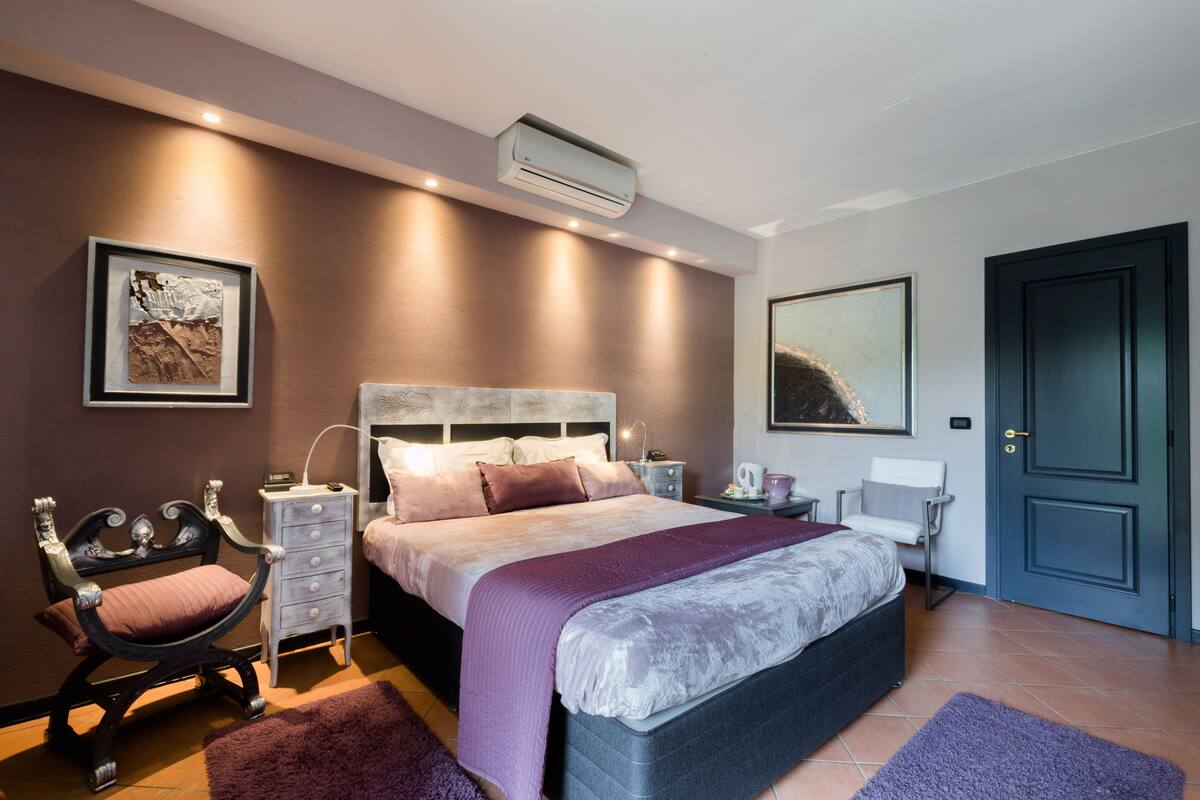 A bedroom with a bed, bedside tables, a chair, an air conditioning unit, and artwork on the walls.