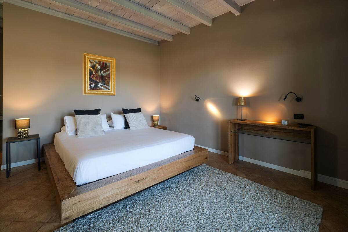 A bedroom with a large bed, a wooden headboard, a desk with a lamp, and a painting on the wall.