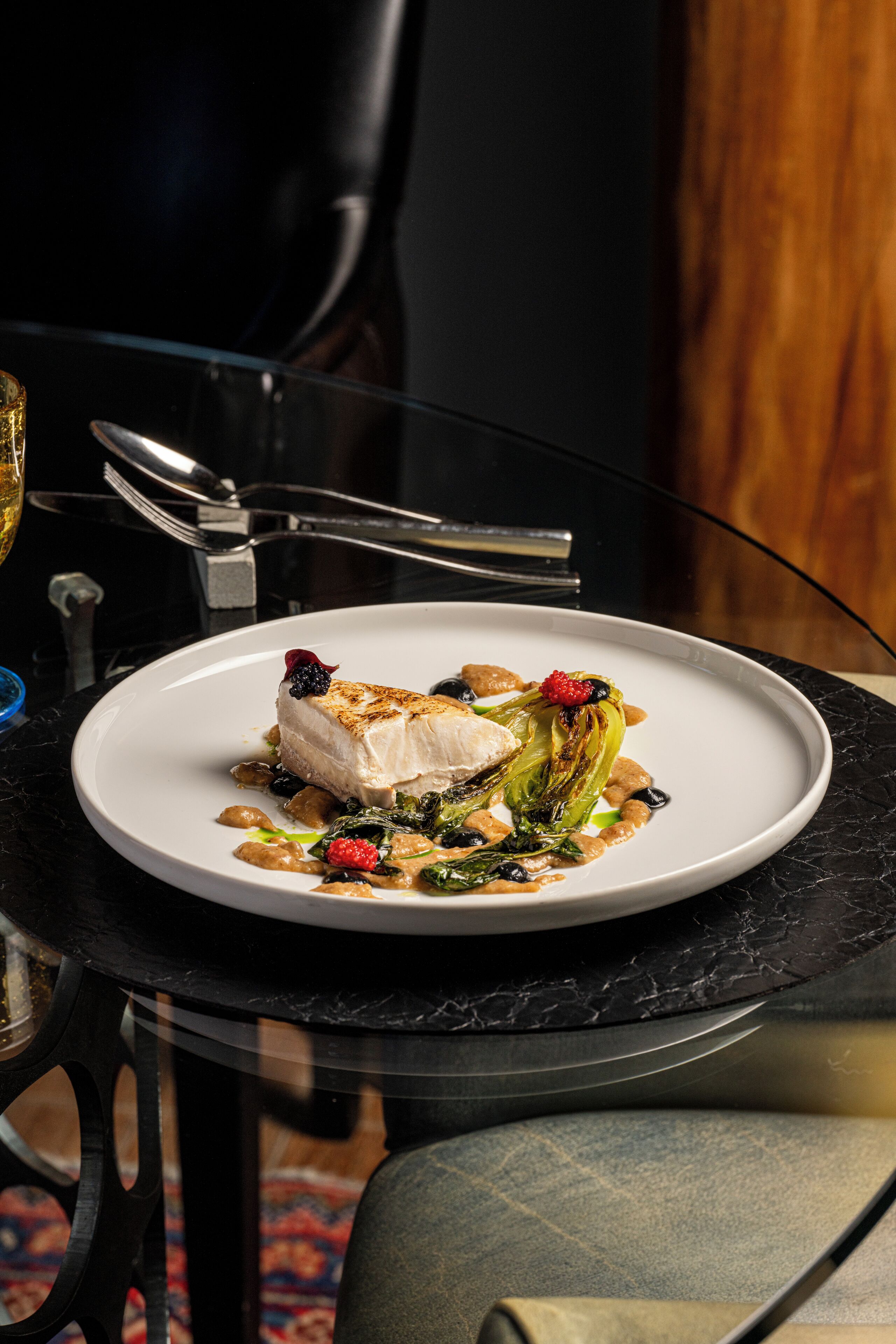 Une assiette blanche contenant un morceau de poisson blanc, des légumes verts et des baies, le tout posé sur une table noire.