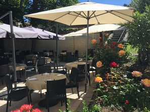 Un área de comedor al aire libre con mesas y sillas bajo sombrillas, rodeada por un jardín con diversas flores.