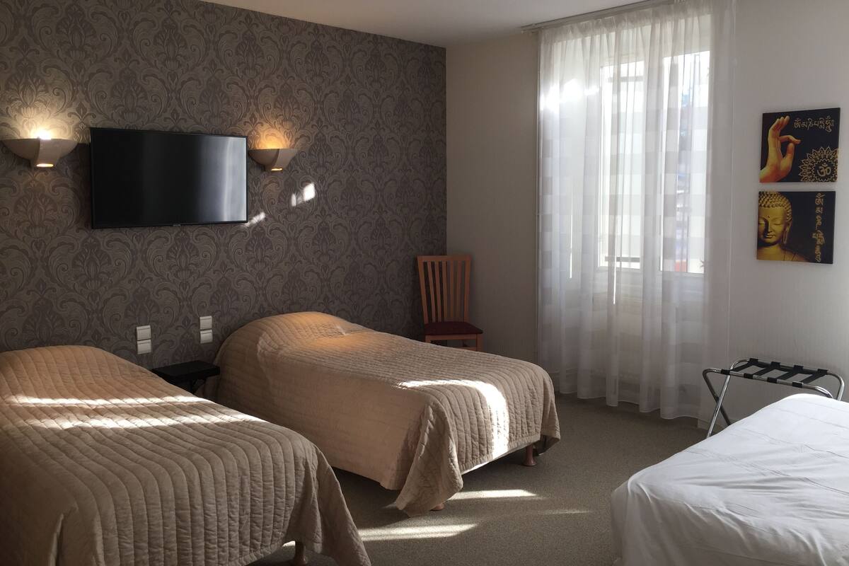 Habitación de hotel con dos camas, un televisor montado en la pared, una silla y una ventana con cortinas translúcidas.