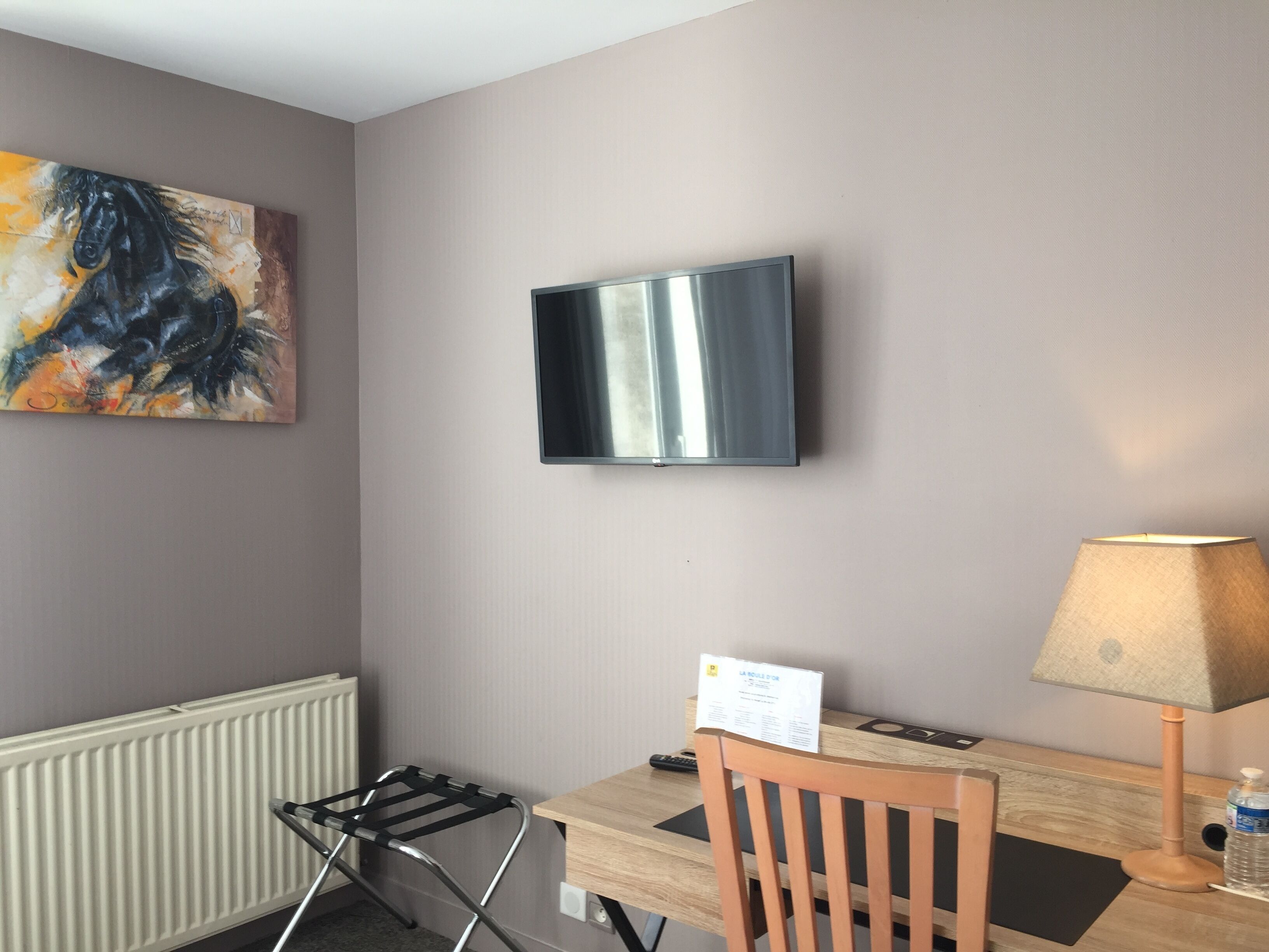 A hotel room with a flat-screen TV mounted on the wall, a wooden desk, a chair, a lamp, and a radiator.
