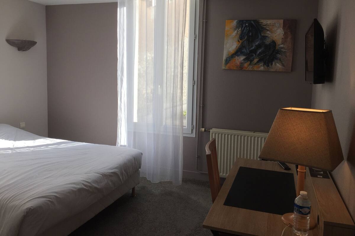 Habitación de hotel con cama, escritorio con lámpara, un cuadro en la pared y ventana con cortinas translúcidas.