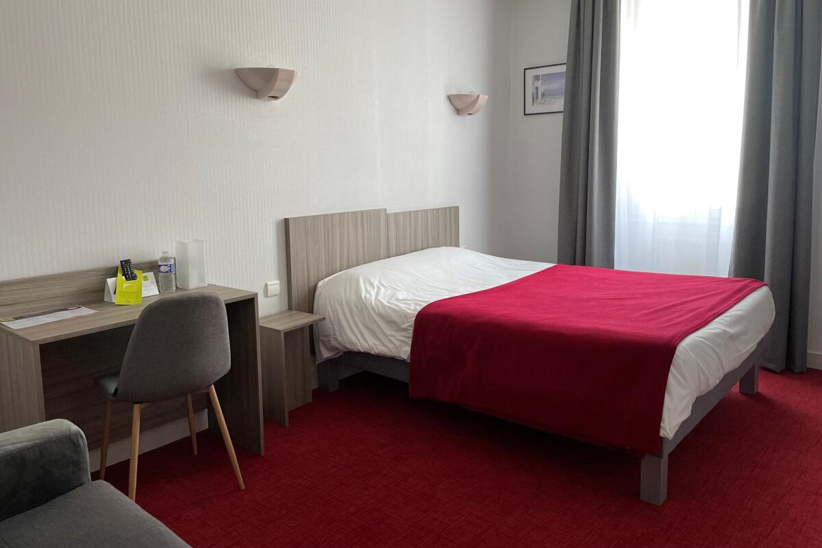 Una habitación de hotel con cama, escritorio, silla, mesita de noche y ventana con cortinas.