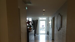 A long, well-lit hallway with a tiled floor, leading to a bright entrance with a glass door.