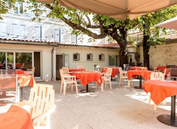 An outdoor patio with tables and chairs, covered by a large umbrella, and a tree providing shade.