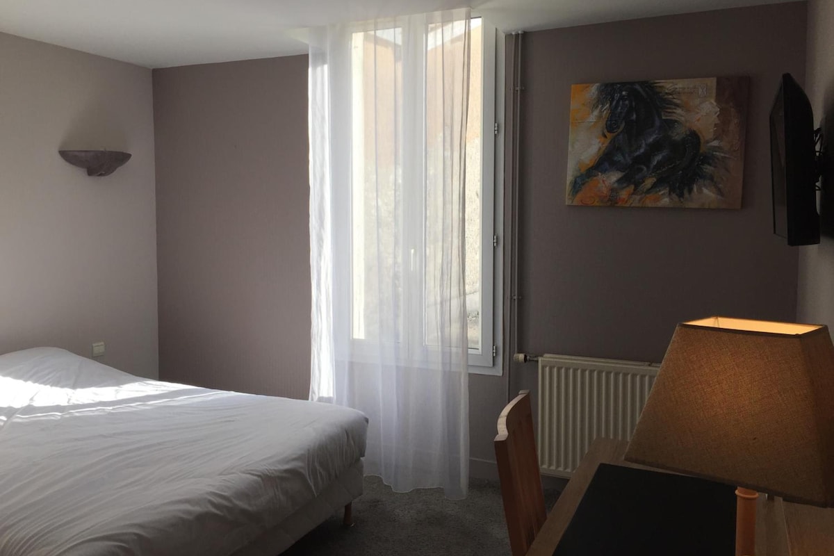 Habitación de hotel con cama, radiador, lámpara, un cuadro y ventana con cortinas translúcidas.