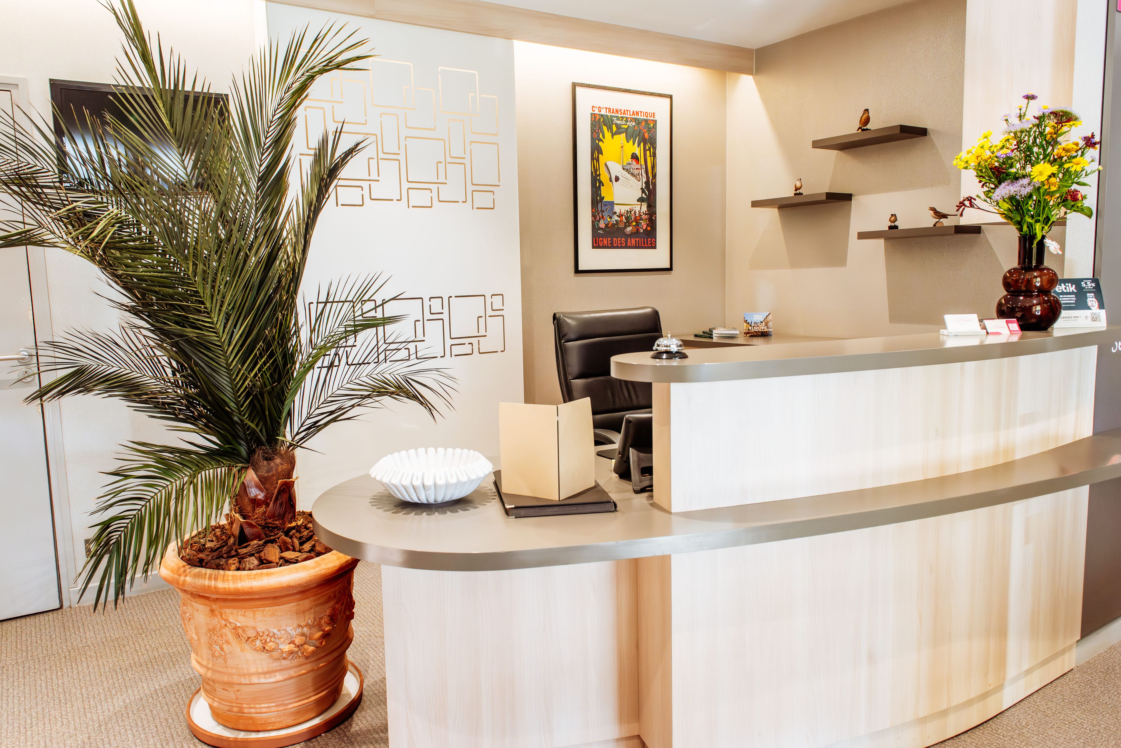 A reception desk with a palm plant, a framed poster, and a vase with flowers.