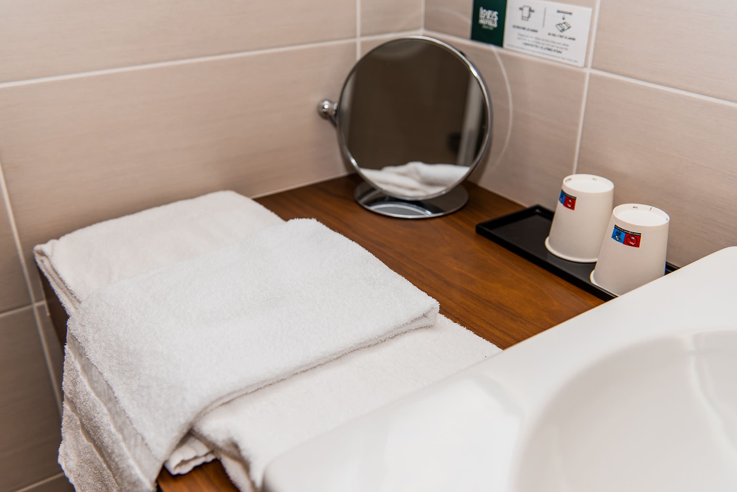 Een badkamer met een houten wastafel, een witte handdoek, een spiegel en twee witte bekers met een rood en blauw logo.