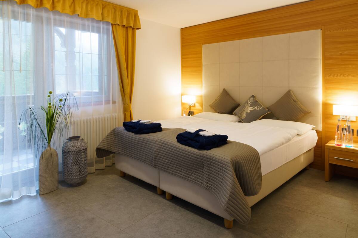 Ein Hotelzimmer mit einem großen Bett, einem Nachttisch mit Lampe, einem Fenster mit durchsichtigen Vorhängen und einer Vase mit Pflanzen.