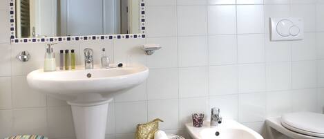 A bathroom with a white sink, bidet, and toilet, a mirror with a decorative frame, and a shelf with toiletries.