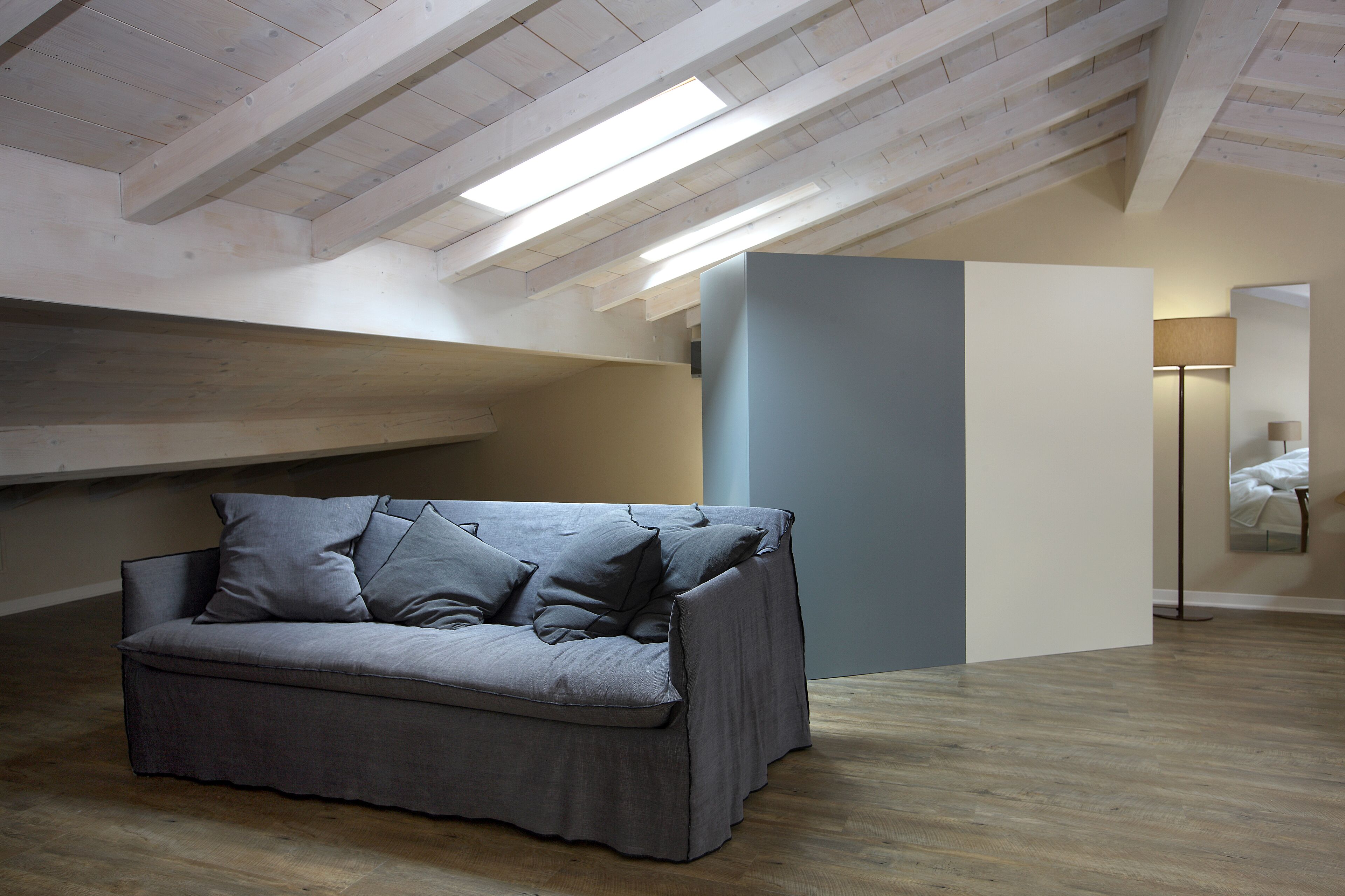 A modern loft bedroom with a grey sofa, wooden beams, and a skylight.