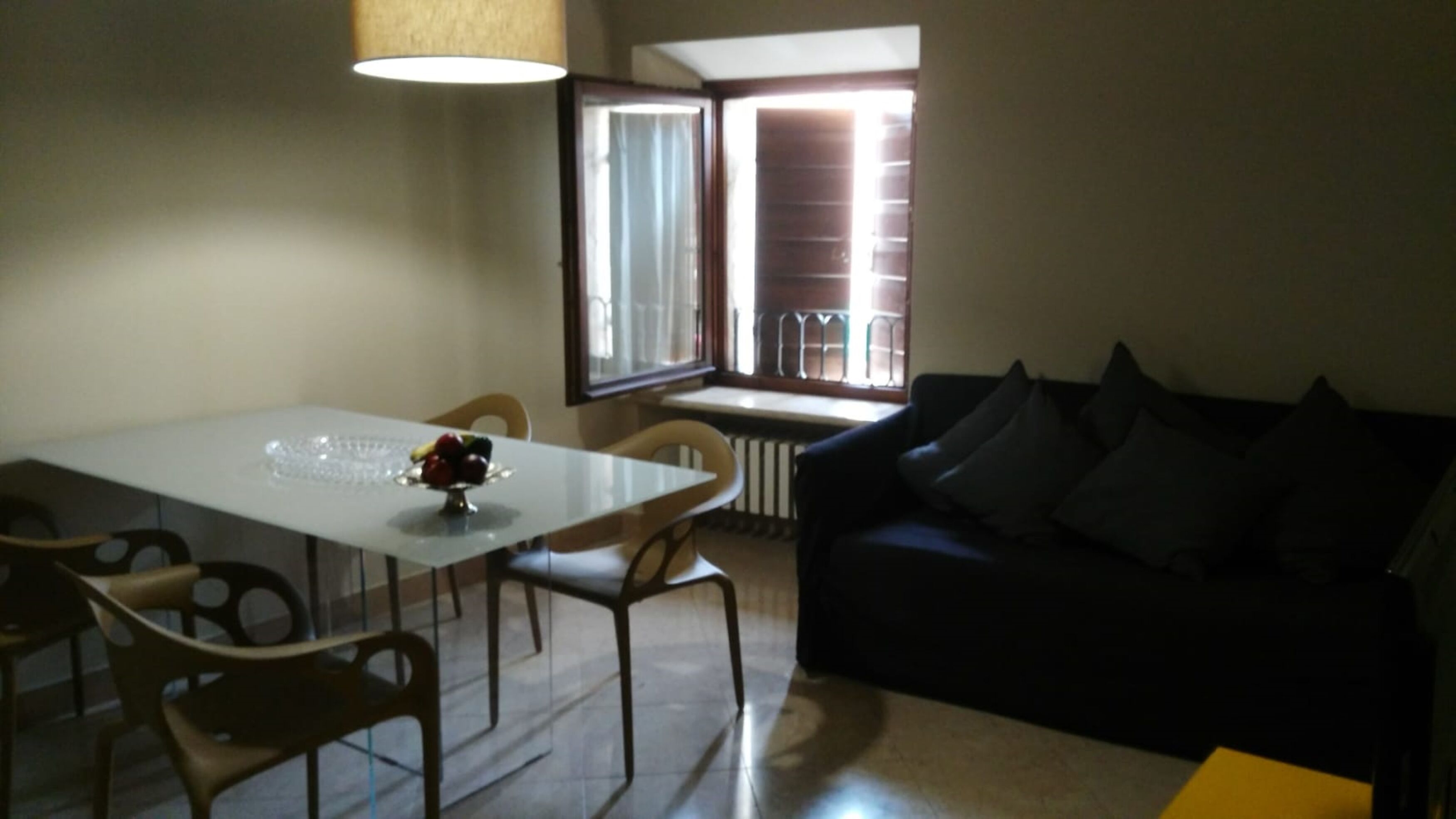 Un comedor con una mesa, sillas y un frutero. Hay un sofá negro con cojines y una ventana con persianas.